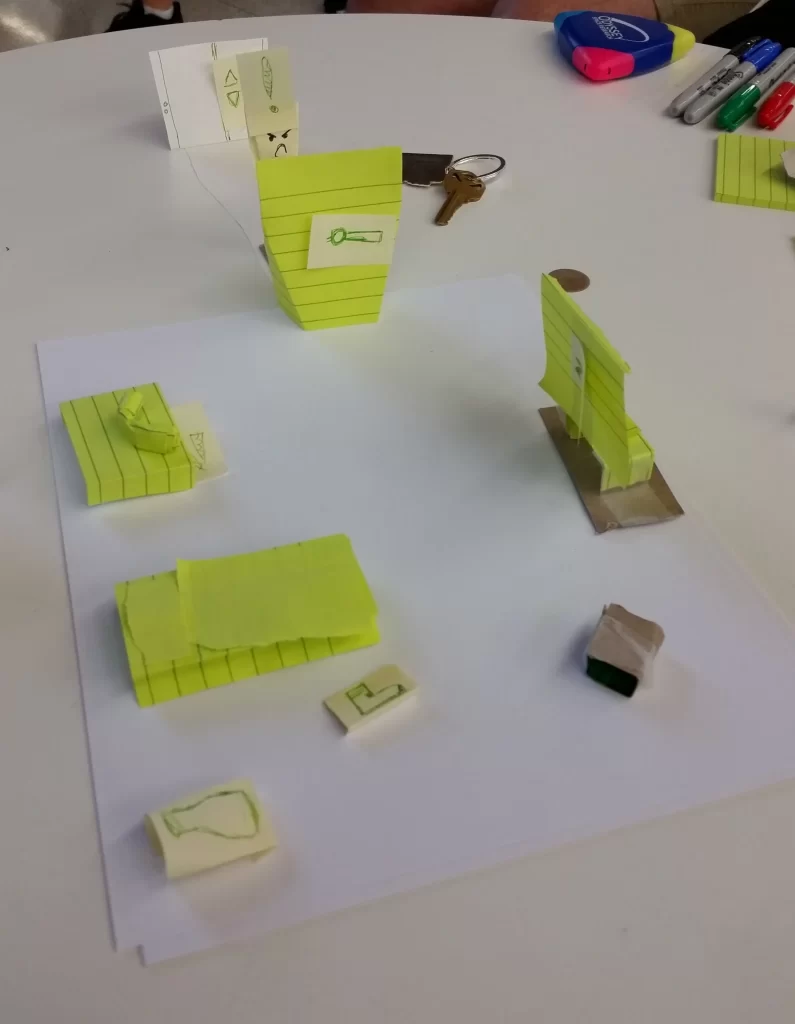 A photo of the paper prototype for the hotel. Different scraps of paper representing the shoebox, hotel furniture, door, shoe pickups, and maid enemy all arranged on a printer paper "room."